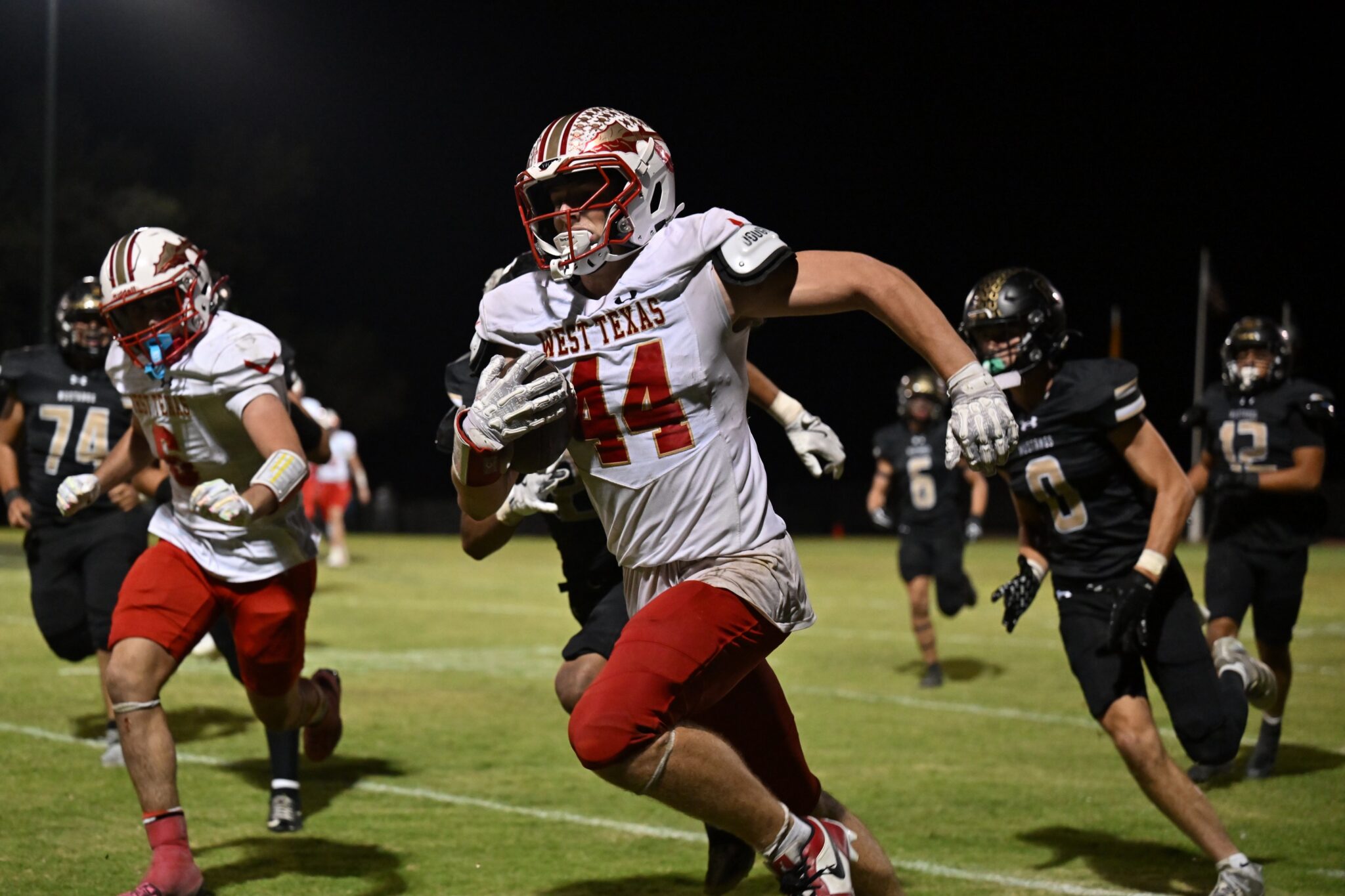 image6 8 | Press Pass Sports West Texas football player wearing white jersey #14 runs with the ball at night, pursued by opposing players on a grassy field.