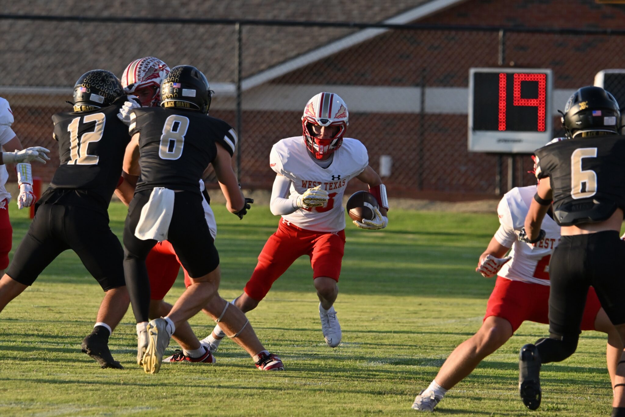 image7 2 | Press Pass Sports Football players in a high school game tackle toward a running back with the ball on a grassy field; scoreboard in the background.