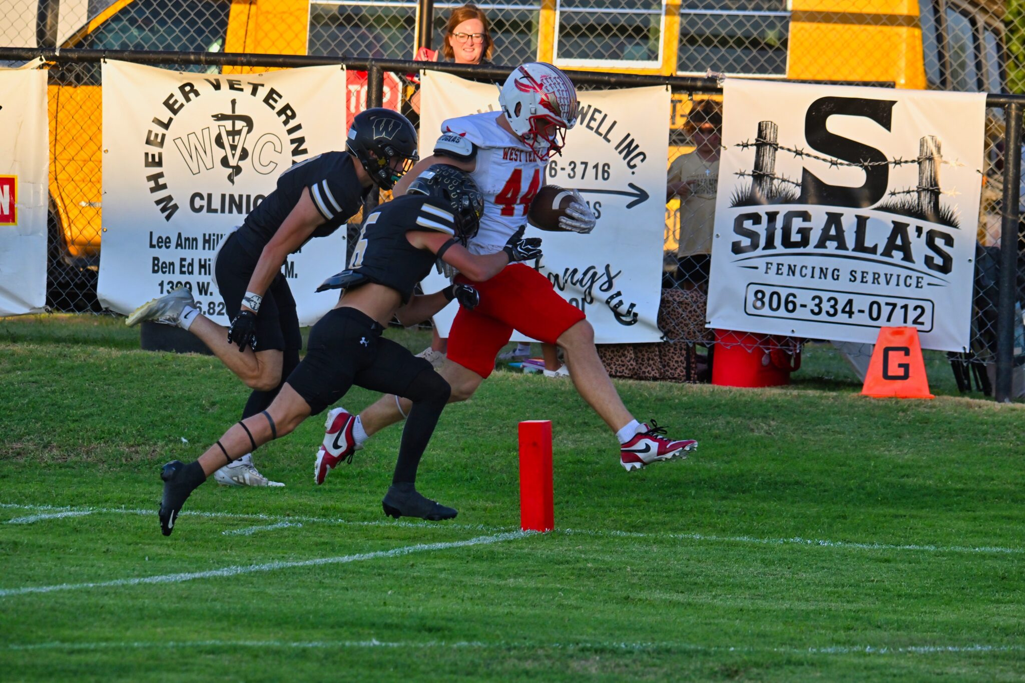 image9 4 | Press Pass Sports Two football players sprint on a grassy field near advertising banners as a referee or spectator watches from behind the fence.