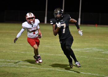 Two football players sprint on a night game; one in white West Texas uniform (#7) chased by a Black Mustangs player (#7).