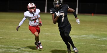 Two football players sprint on a night game; one in white West Texas uniform (#7) chased by a Black Mustangs player (#7).