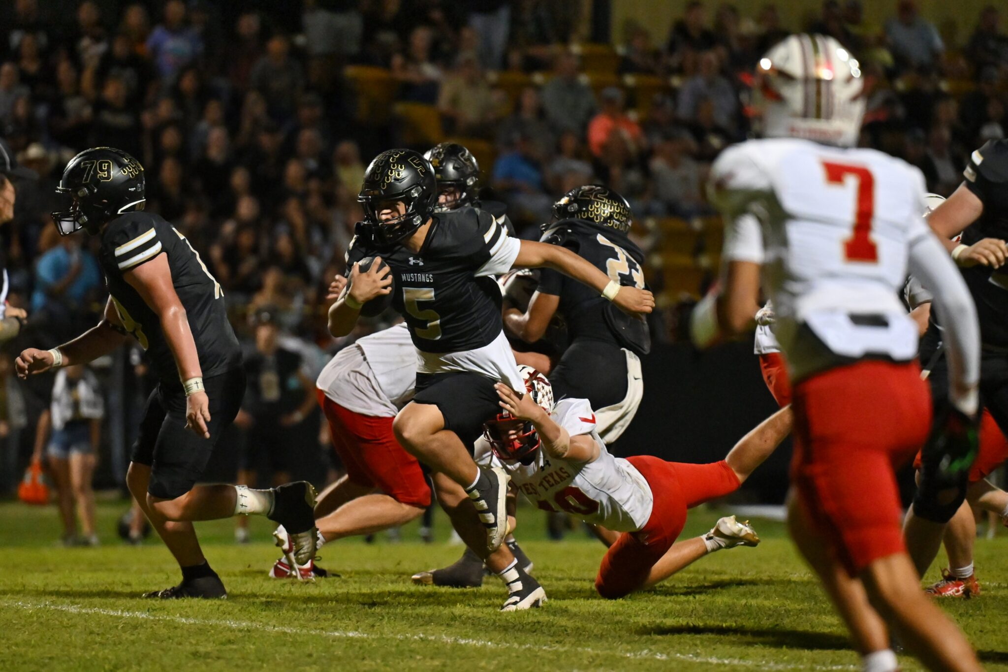 image10 2 | Press Pass Sports American football players in black uniforms chase the ball carrier as a white-jersey defender reaches in during a nighttime game.