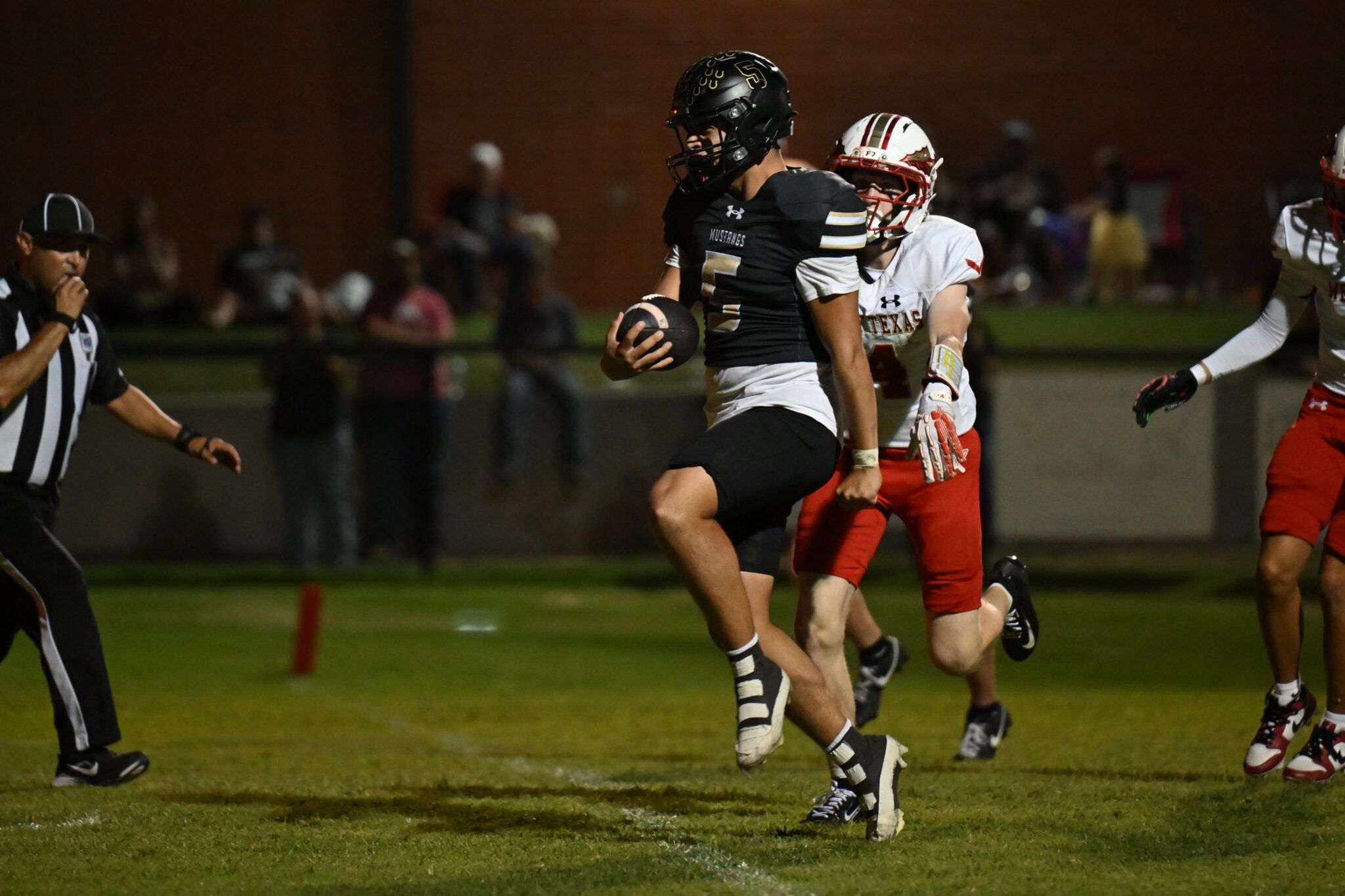 image11 1 | Press Pass Sports Football player in a black Mustangs jersey runs with the ball as a defender in white and red pursues him on a night game field.