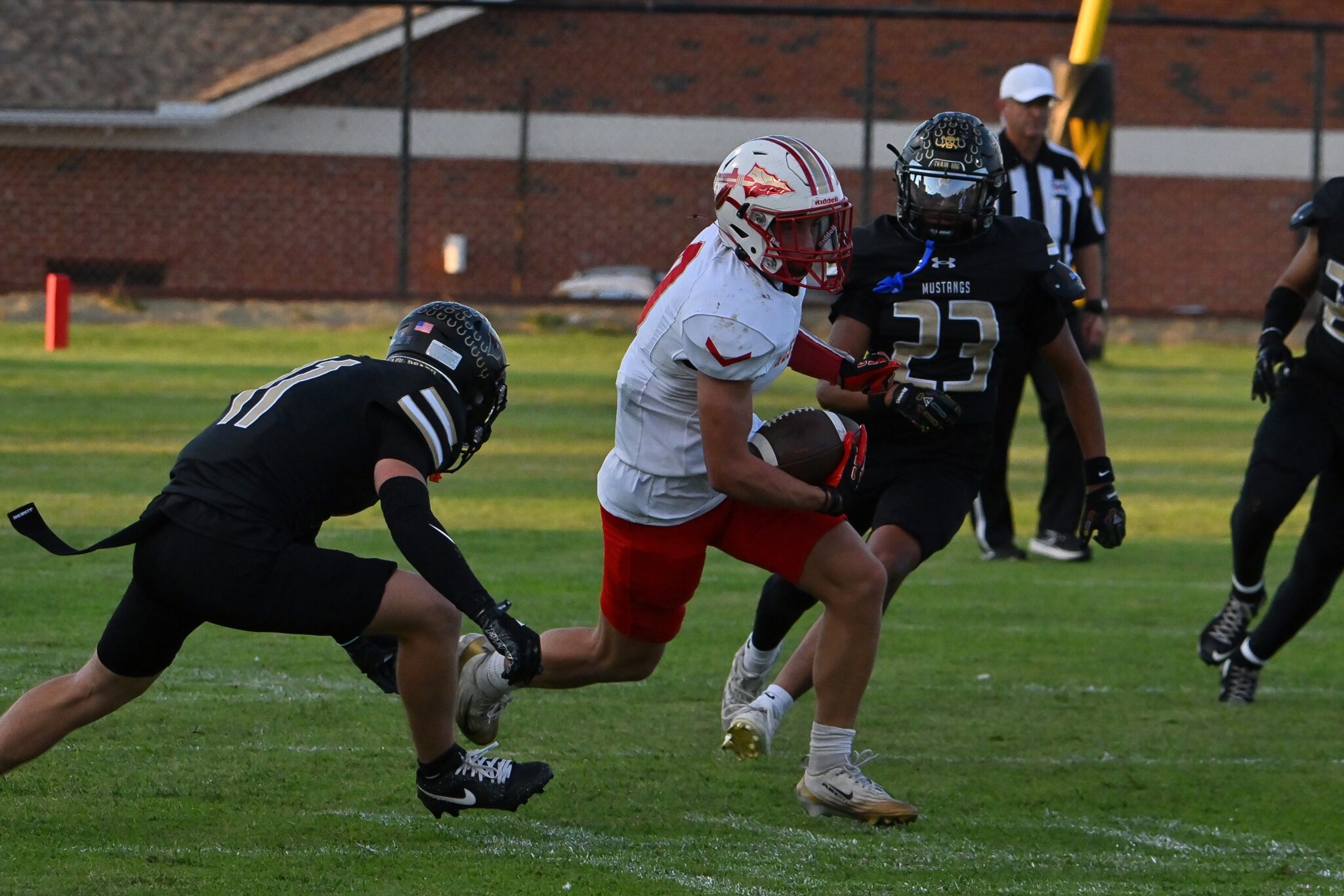 image11 | Press Pass Sports Football action: a white-jersey player carrying the ball pursued by a black-uniform defender on a grassy field with a brick backdrop.