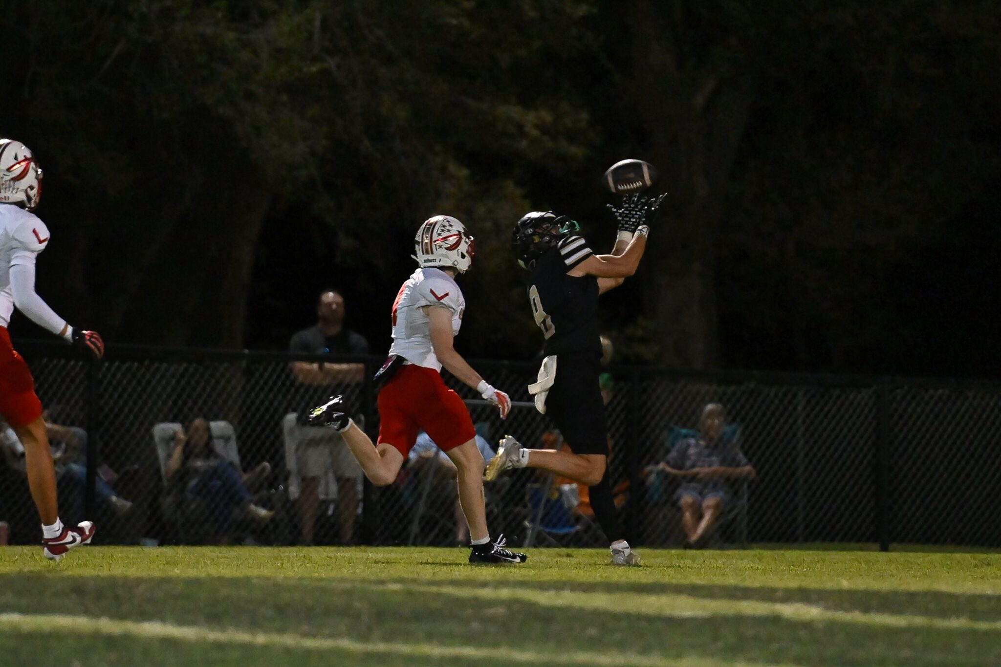 image12 | Press Pass Sports Football players jump for a catch near the sideline during a night game; one in black with outstretched arms reaches for the ball while others watch behind a fence.