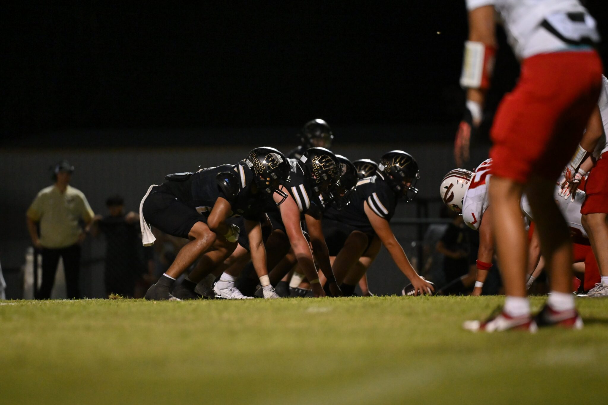 image14 | Press Pass Sports Night game football: offensive linemen in black crouch at the line of scrimmage, facing red-clad defenders on a grassy field.