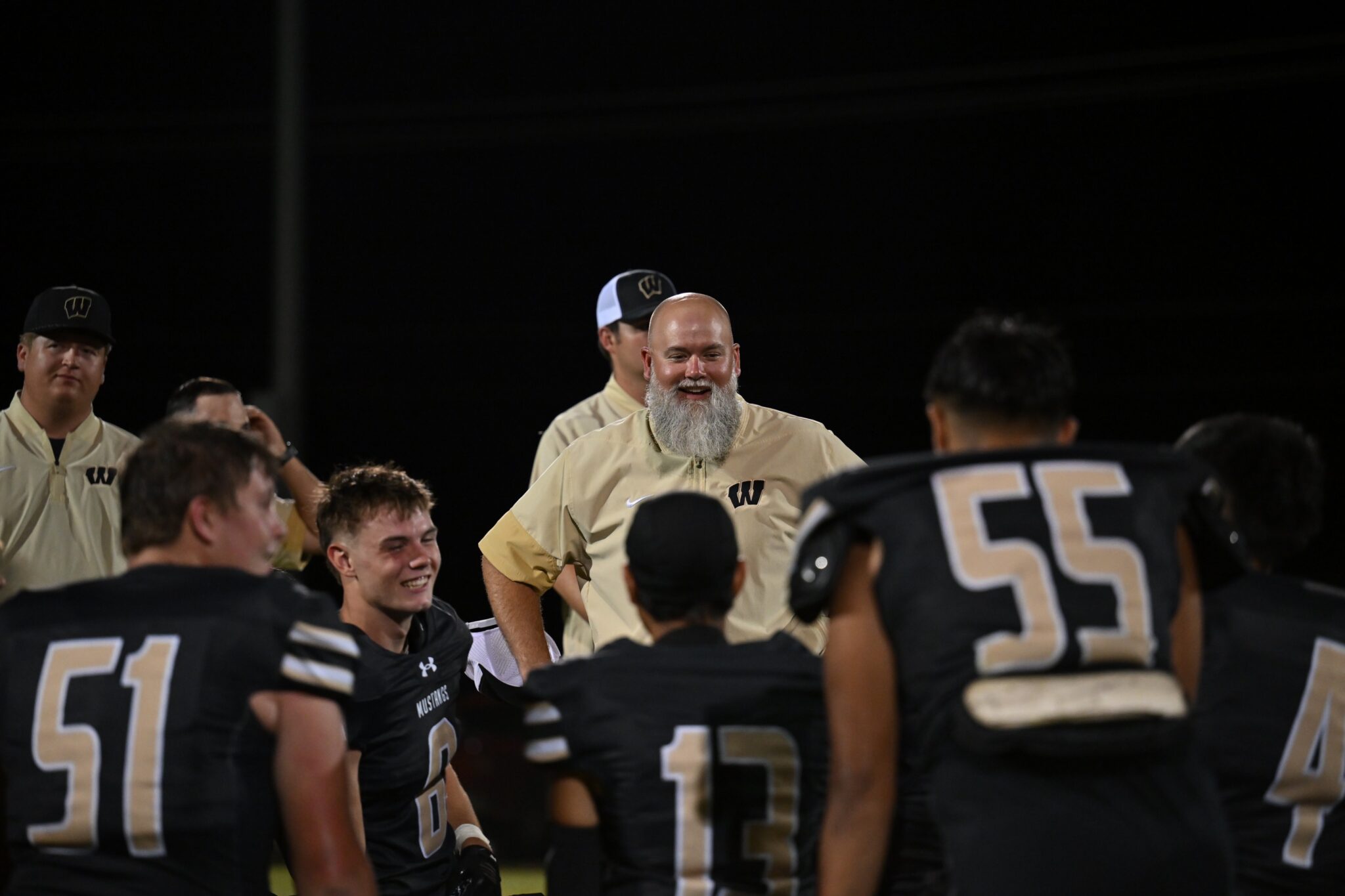 image15 | Press Pass Sports Group of football players in beige jackets and black jerseys stand together on a dark field at night; central figure has a gray beard and smiles.