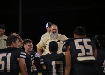 image15 | Press Pass Sports Group of football players in beige jackets and black jerseys stand together on a dark field at night; central figure has a gray beard and smiles.