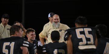 Group of football players in beige jackets and black jerseys stand together on a dark field at night; central figure has a gray beard and smiles.