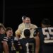 image15 | Press Pass Sports Group of football players in beige jackets and black jerseys stand together on a dark field at night; central figure has a gray beard and smiles.