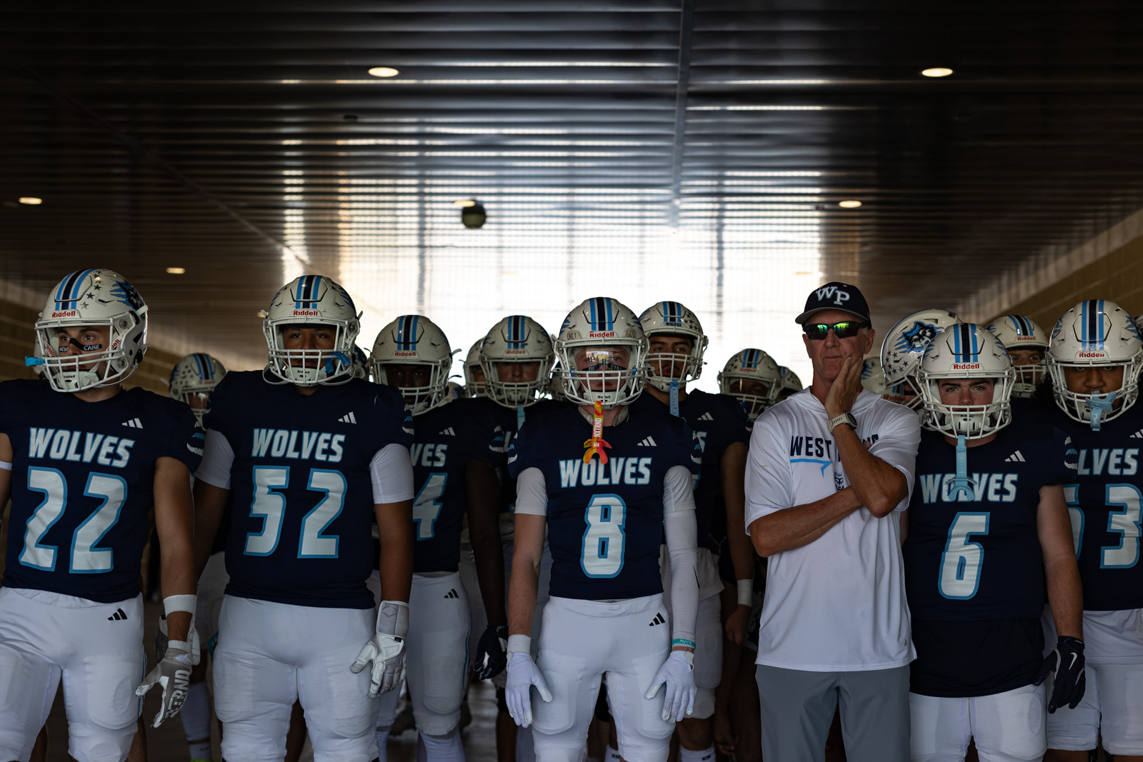 LHS-WPHS-FB-1 | Press Pass Sports Wolves football players in navy jerseys and white pants stand in a tunnel, ready for a game, with a coach on the right.