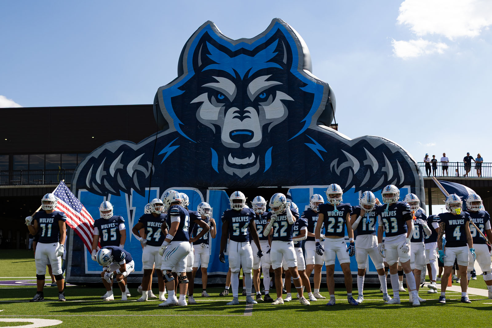 LHS-WPHS-FB-2 | Press Pass Sports College football team stands in front of a large blue wolf inflatable logo on the field, with an American flag nearby.