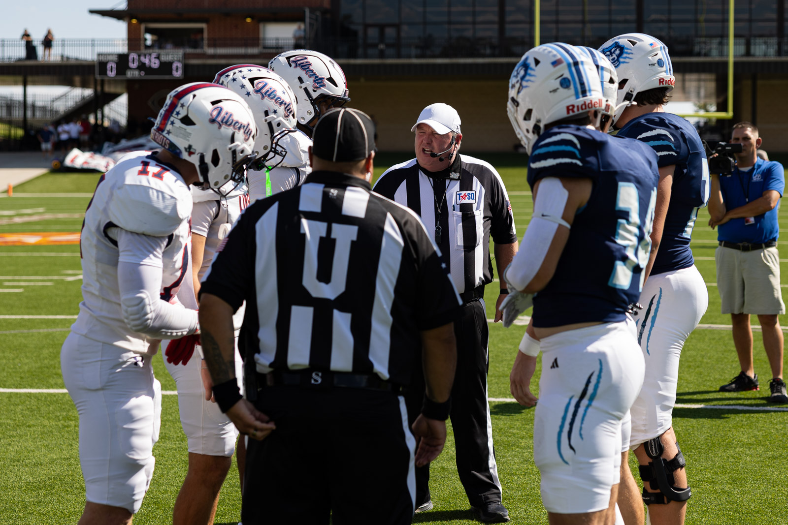 LHS-WPHS-FB-3 | Press Pass Sports Referee with headset talks to two football teams lined up on the sunny field before kickoff, white and blue uniforms facing each other on the line of scrimmage.