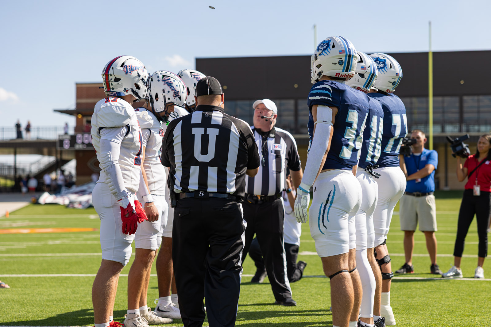 LHS-WPHS-FB-6 | Press Pass Sports Football captains from white and blue teams face off at midfield as referees brief them during a coin toss on a sunny field.