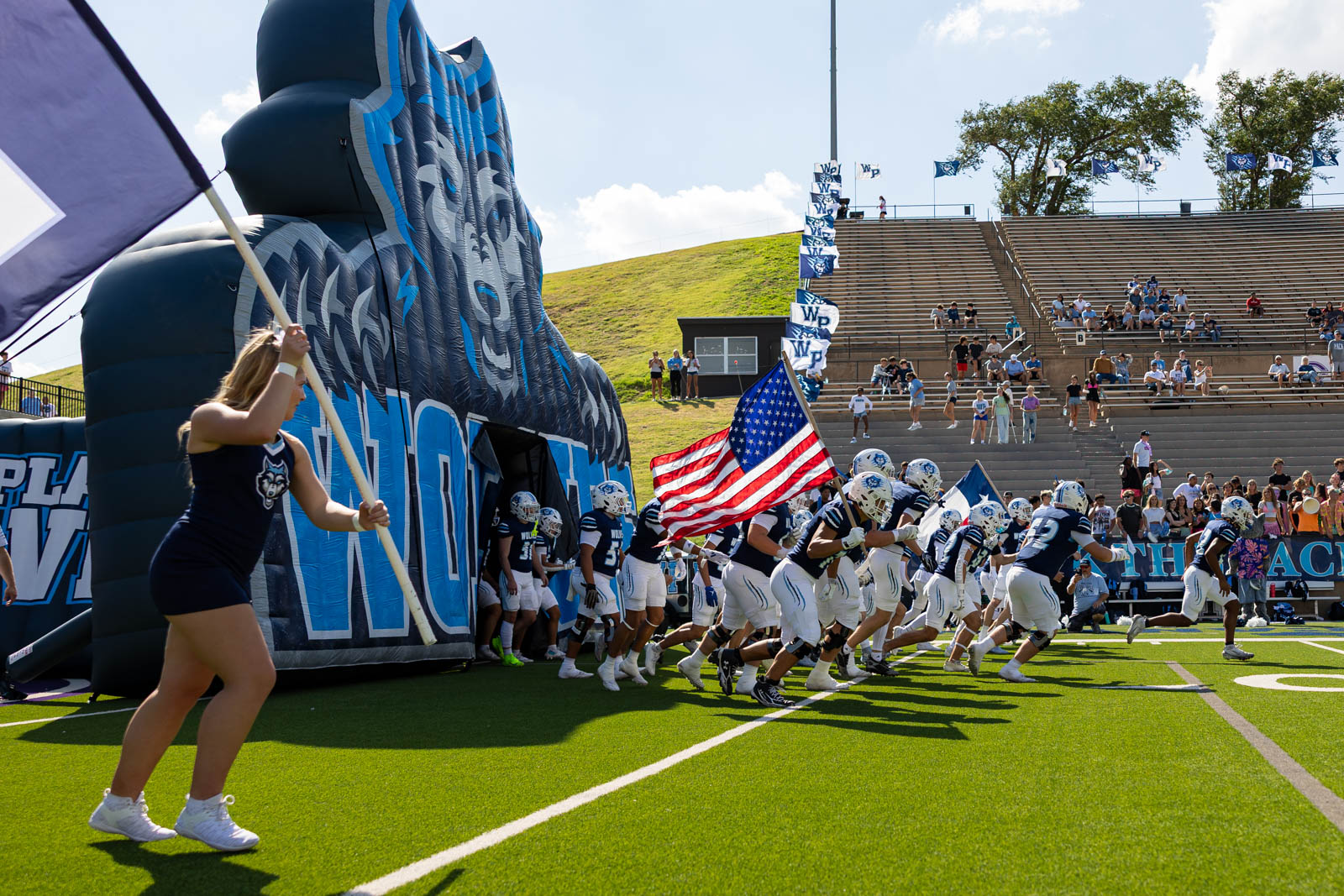 LHS-WPHS-FB-11 | Press Pass Sports Football team runs onto the field through a blue inflatable tunnel while a cheerleader with a flag leads them.