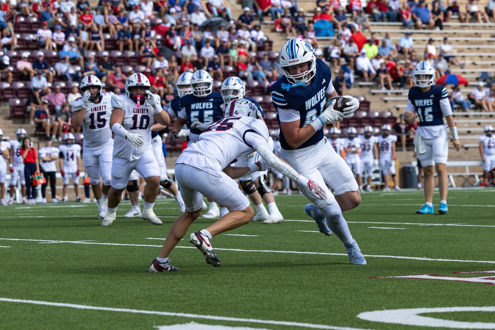 LHS-WPHS-FB-17 | Press Pass Sports Wolves running back in navy jersey carries the ball while a defender in white dives in from the side on a green football field, as supporters watch from the stands.