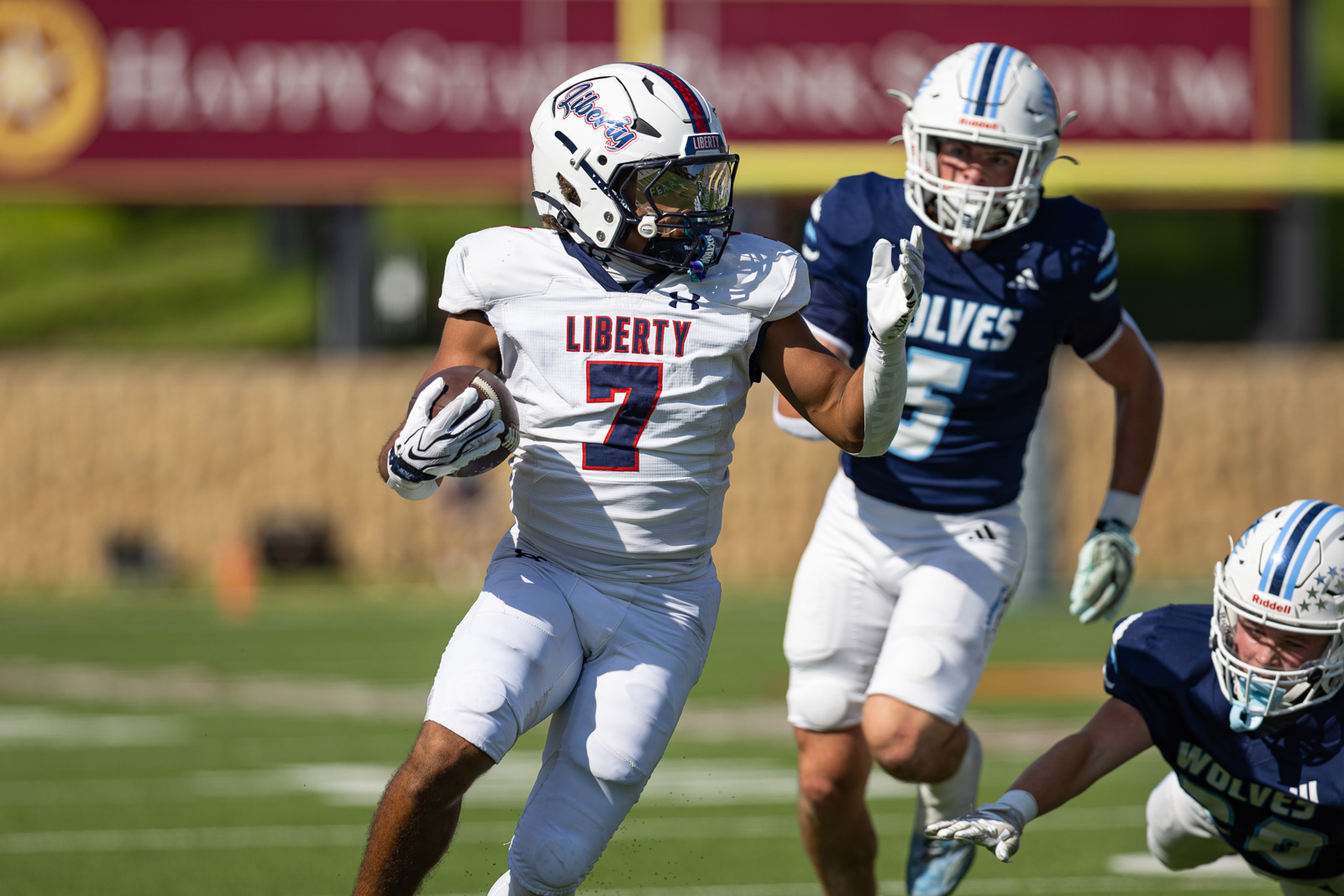 LHS-WPHS-FB-20 | Press Pass Sports Liberty player in a white jersey #7 runs with the football as a defender in blue pursues him down the field.