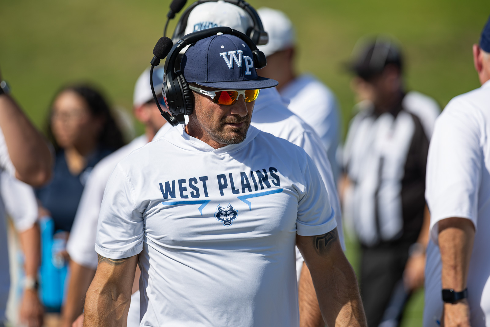 LHS-WPHS-FB-21 | Press Pass Sports Coach wearing a headset and sunglasses in a white West Plains shirt on a sunny sports field.
