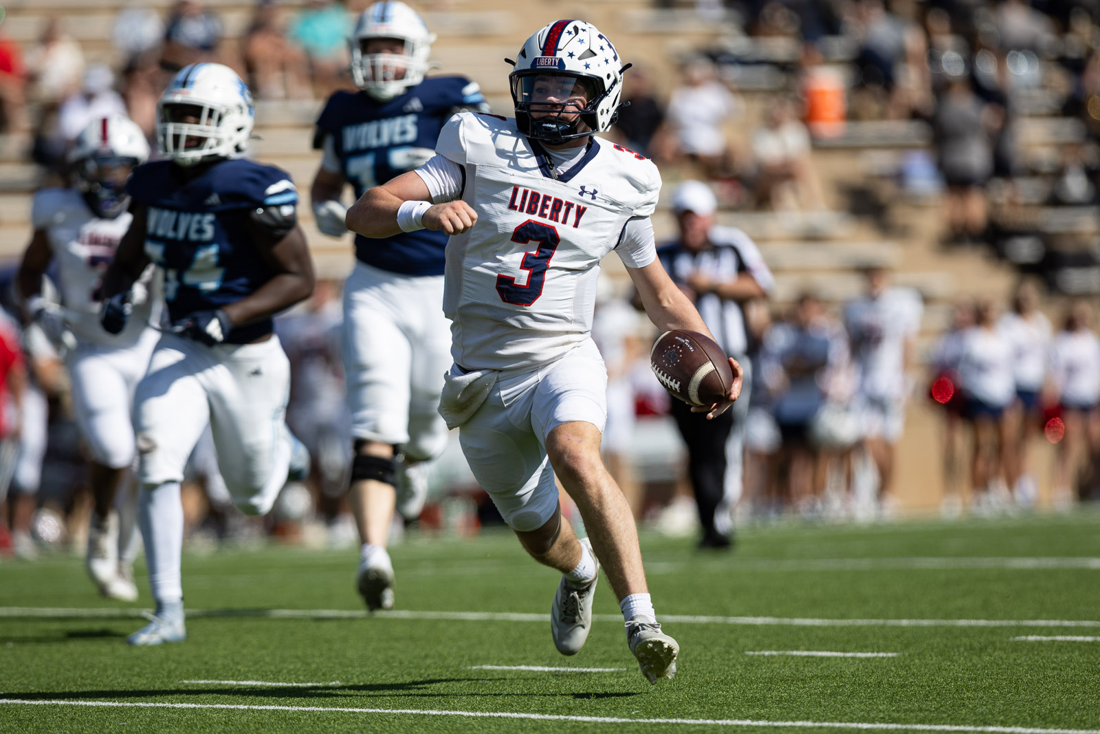 LHS-WPHS-FB-24 | Press Pass Sports Liberty quarterback number 3 sprints with the football during a game, pursued by opponents in blue uniforms on a sunny field.