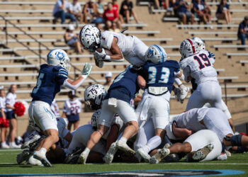 LHS-WPHS-FB-25 | Press Pass Sports White-team ball carrier jumps over blue-team defenders as players pile up on the football field during a game.