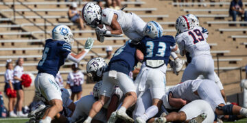 White-team ball carrier jumps over blue-team defenders as players pile up on the football field during a game.