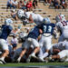 White-team ball carrier jumps over blue-team defenders as players pile up on the football field during a game.