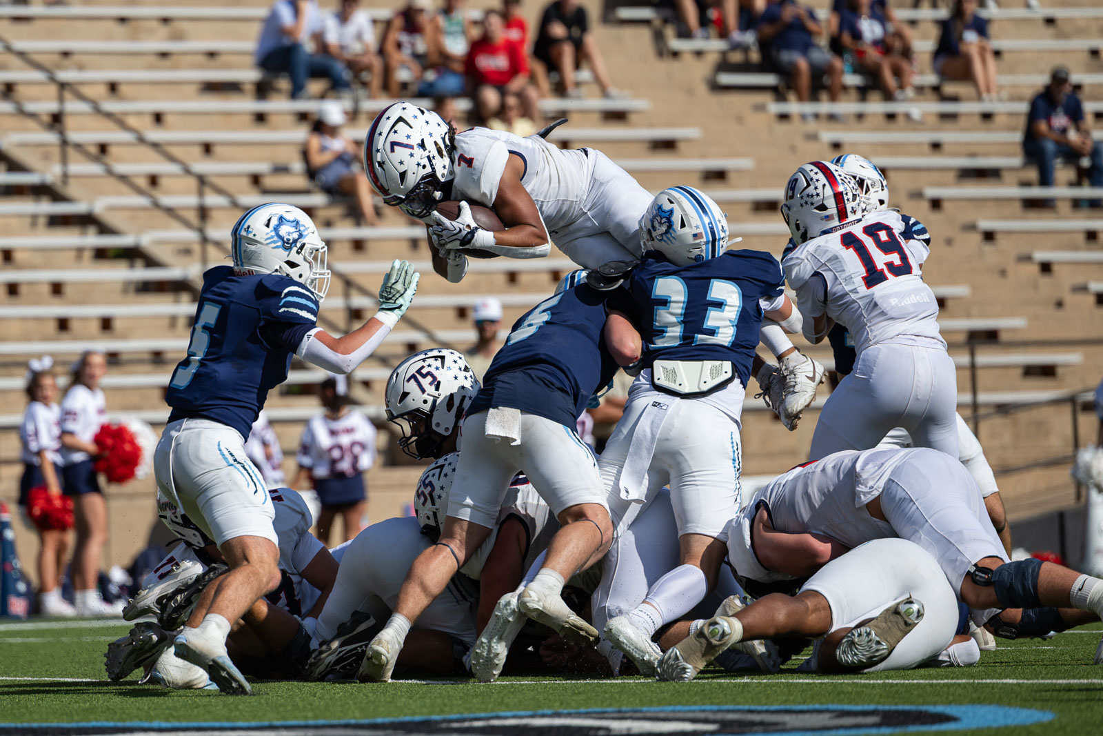 LHS-WPHS-FB-25 | Press Pass Sports White-team ball carrier jumps over blue-team defenders as players pile up on the football field during a game.