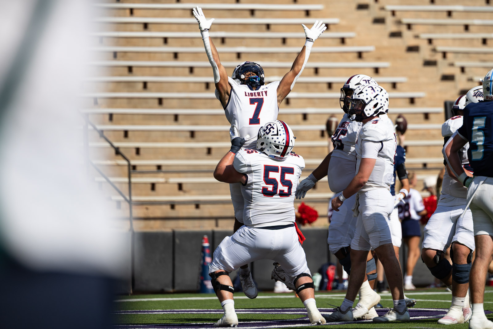 LHS-WPHS-FB-26 | Press Pass Sports Liberty football player #7 jumps with arms raised in celebration as teammates mob him on a sunny field.