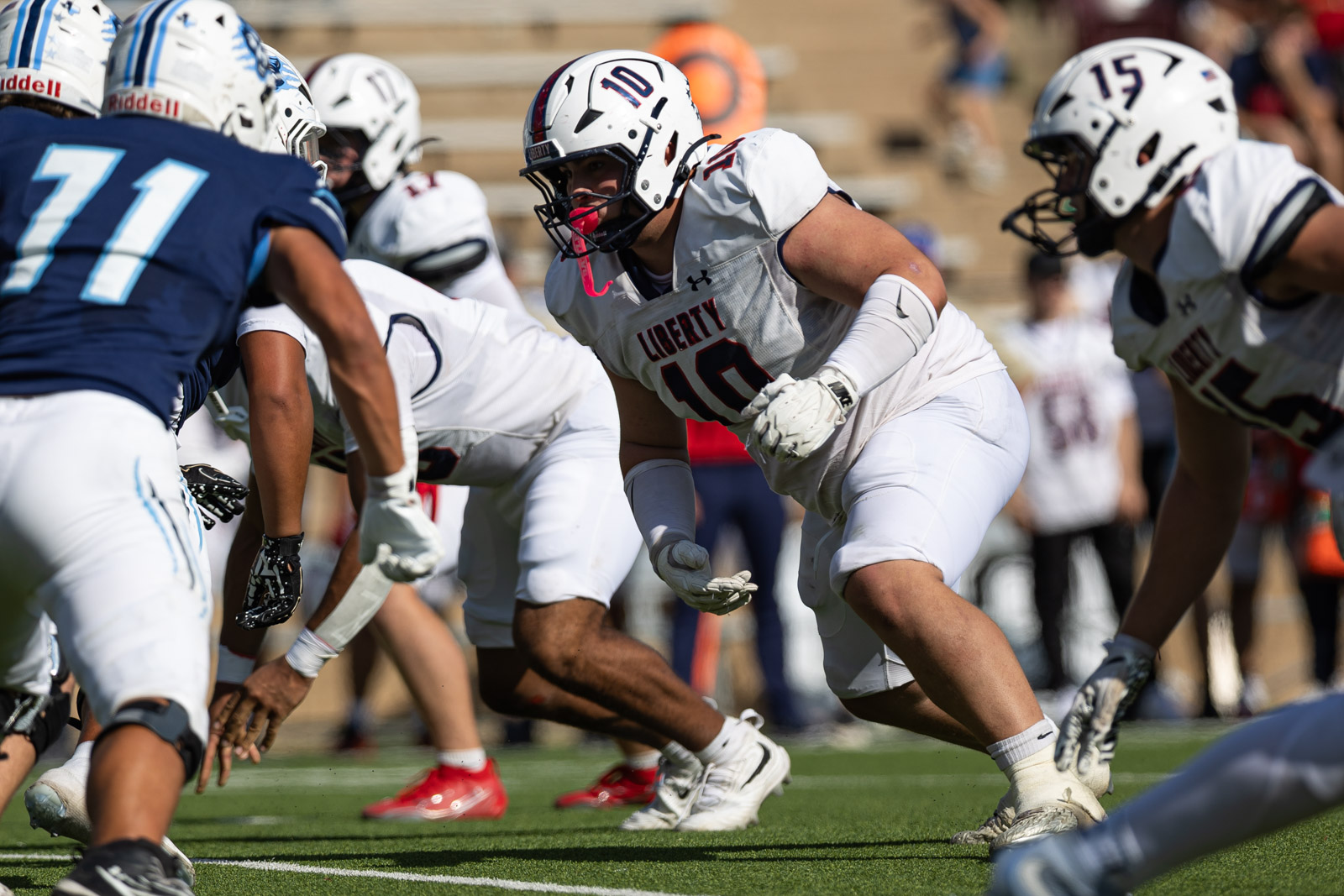 LHS-WPHS-FB-27 | Press Pass Sports Football players in white Libert y uniforms collide at the line of scrimmage during a daytime game, with a player wearing number 10 in focus and a pink mouthguard visible behind his facemask.