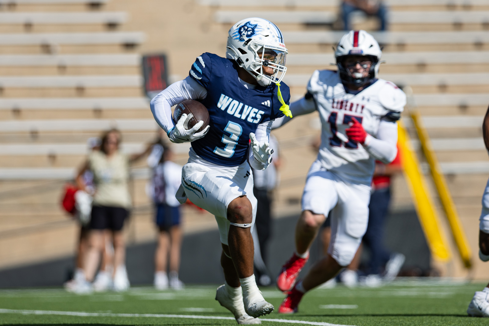 LHS-WPHS-FB-28 | Press Pass Sports Football player in a navy Wolves jersey (number 3) runs with the ball as a defender in white closes in on a green turf field with stands in the background.