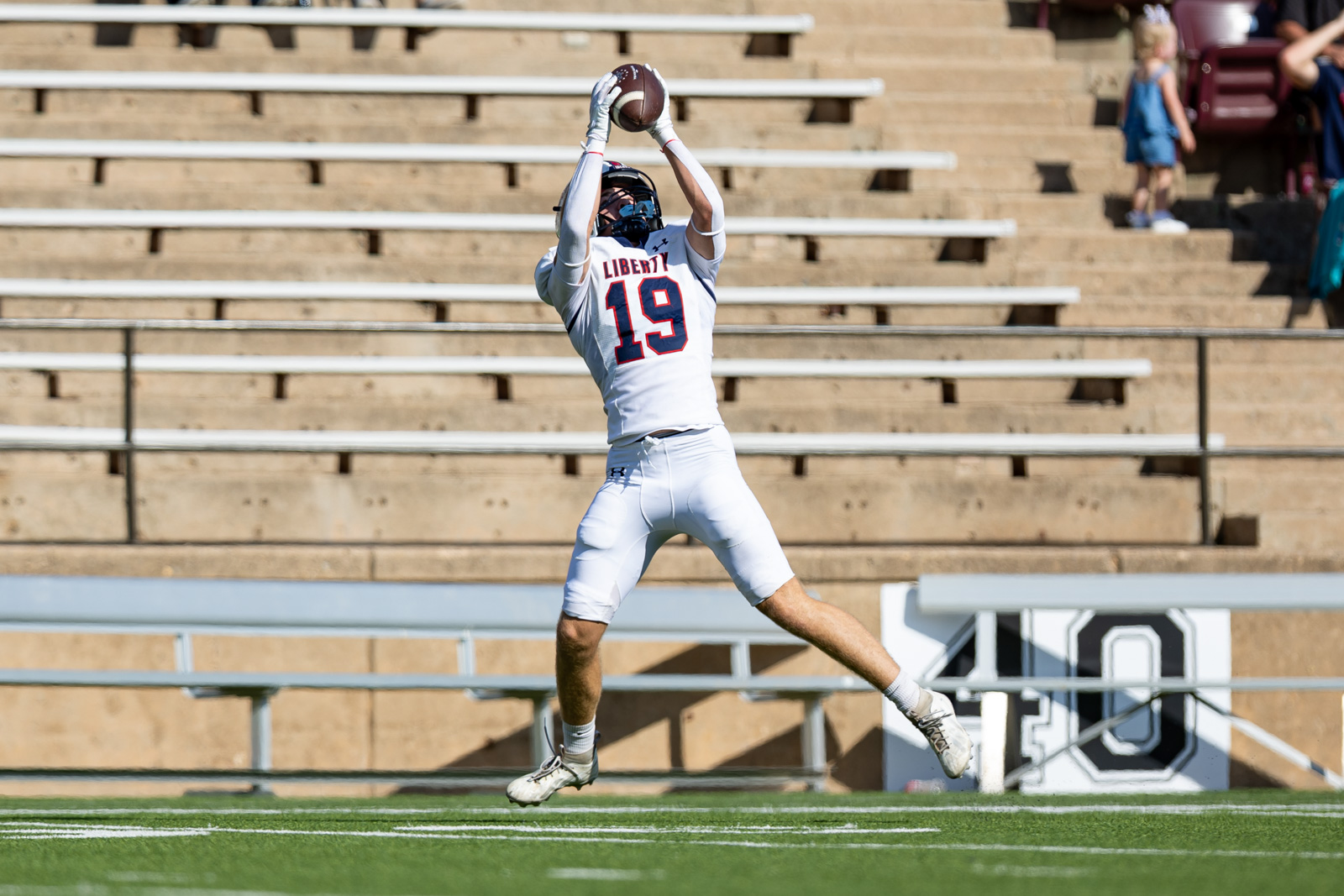 LHS-WPHS-FB-30 | Press Pass Sports Football player in a white Liberty jersey #19 leaping to catch a high pass on a green field with empty bleachers behind him.