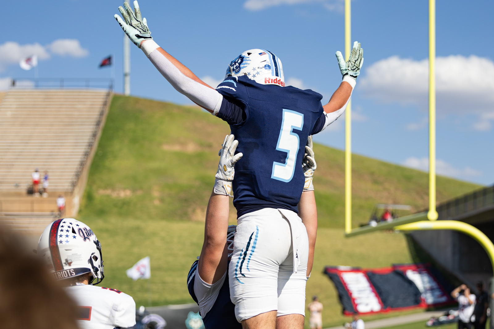 LHS-WPHS-FB-36 | Press Pass Sports Football players celebrate a touchdown as one lifts another above the field, number 5 in blue jersey visible against a clear sky.