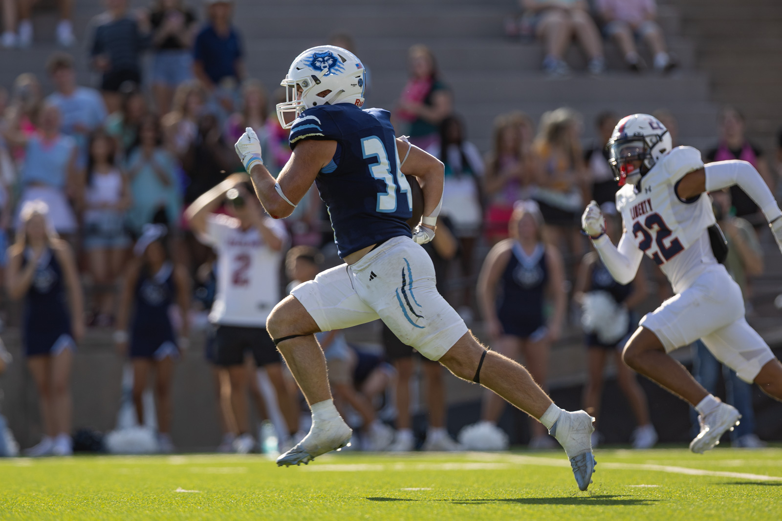 LHS-WPHS-FB-42 | Press Pass Sports Football players sprint down the field during a game, navy blue and white uniforms, crowd in the stands behind them.