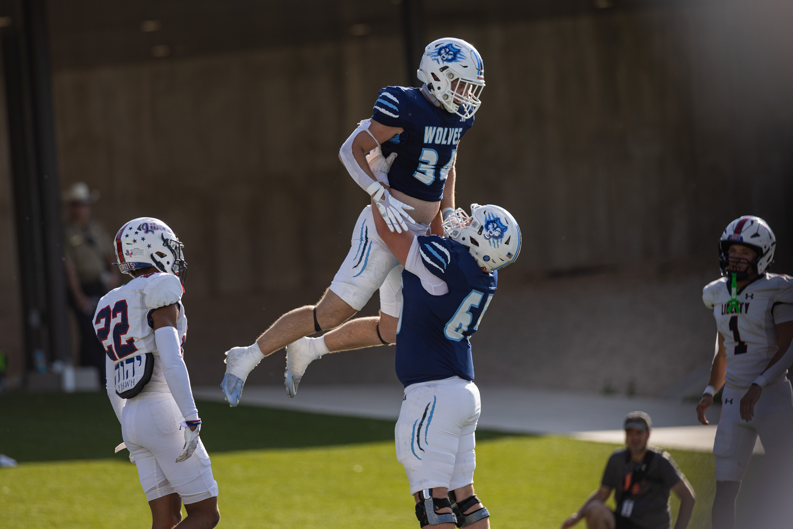 LHS-WPHS-FB-44 | Press Pass Sports Two football players in blue jerseys lift a teammate into the air during a game on a green field. Other players watch nearby.
