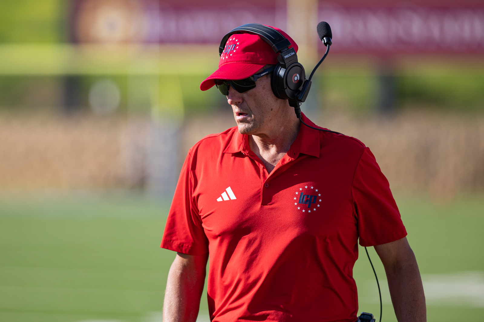 LHS-WPHS-FB-45 | Press Pass Sports Coach wearing a red polo shirt, cap, and sunglasses with a headset, standing on a sunny sports field.