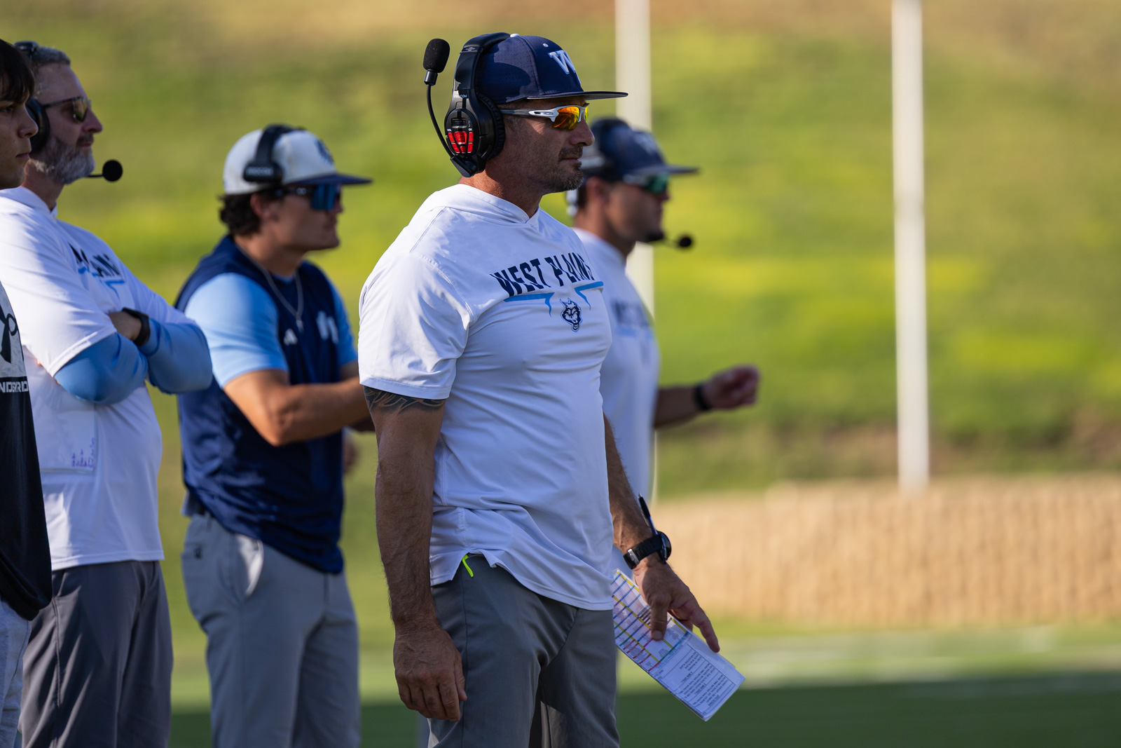 LHS-WPHS-FB-47 | Press Pass Sports Coach in white West Fargo shirt with headset stands on sideline holding a play sheet as others watch on a sunny field.