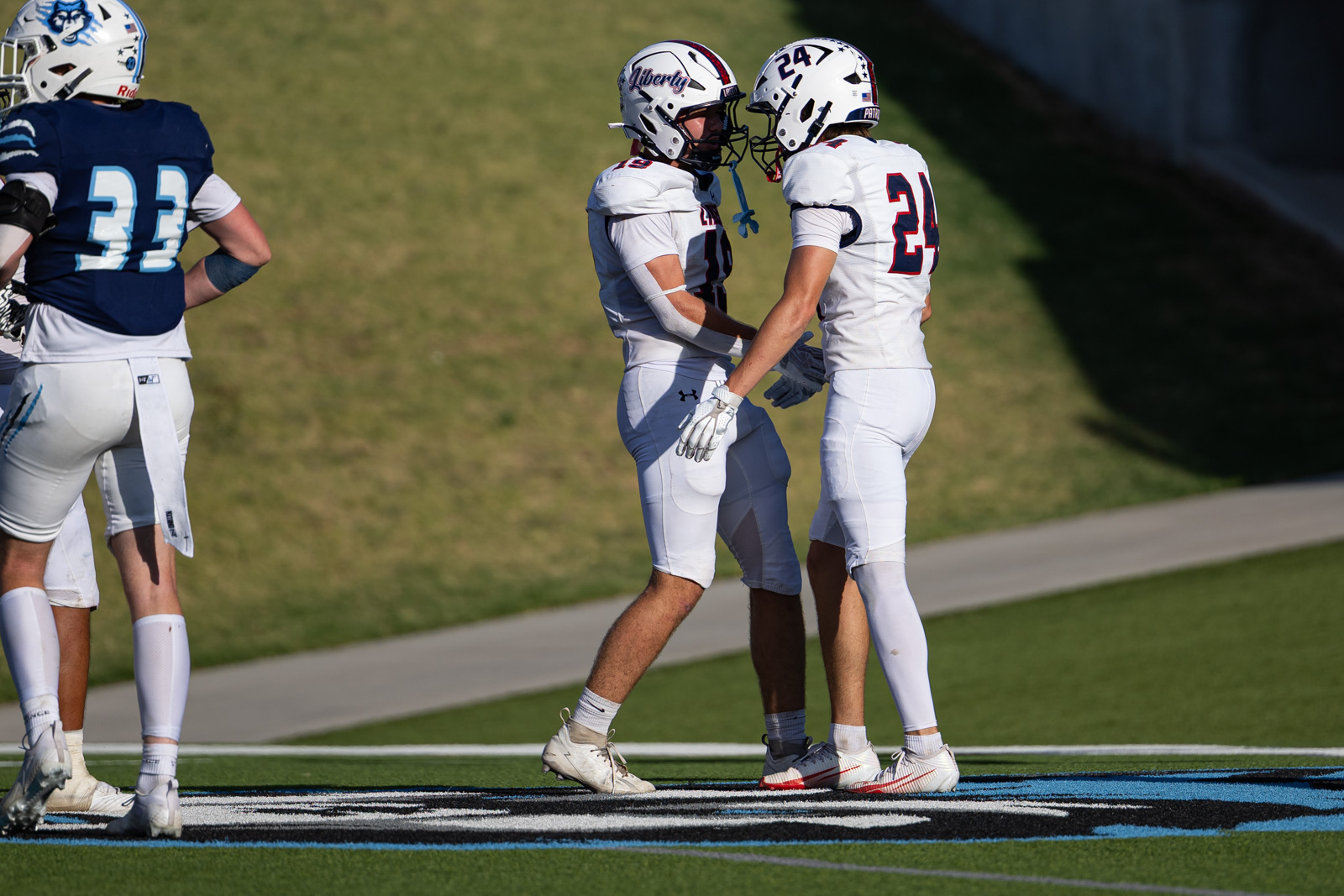 LHS-WPHS-FB-48 | Press Pass Sports Two football players in white uniforms face off and touch helmets on a grassy field, with a blue-jerseyed player nearby.
