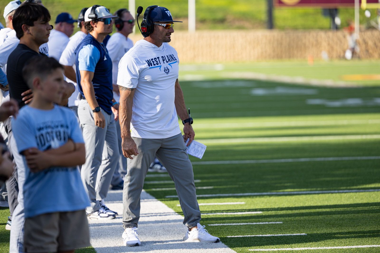 LHS-WPHS-FB-49 | Press Pass Sports Coach wearing a West Plains shirt and headset, holding a play sheet, standing with players on the football sideline.
