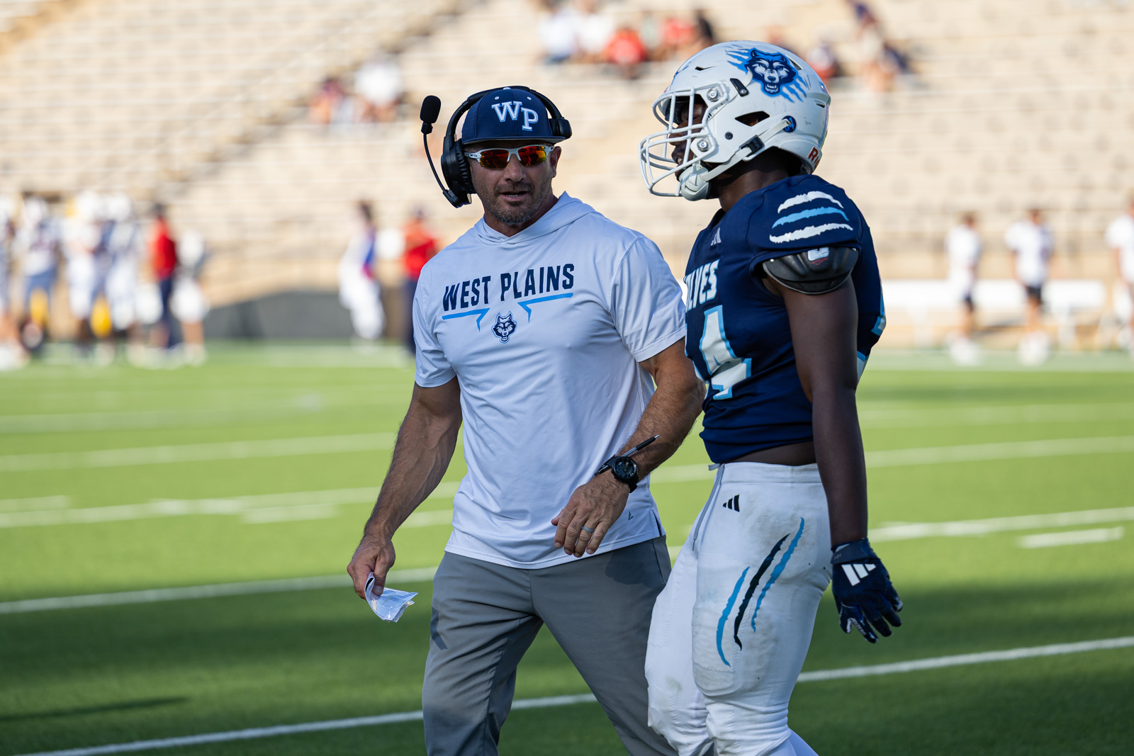 LHS-WPHS-FB-50 | Press Pass Sports Coach with headset and 'West Plains' shirt talks to a football player in a blue uniform on a sunny field.