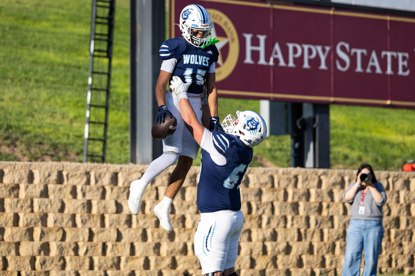 LHS-WPHS-FB-54 | Press Pass Sports Two football players in navy uniforms leap in the air; one catches or guards the ball while the other reaches up, with a maroon sign reading 'Happy State' in the background.