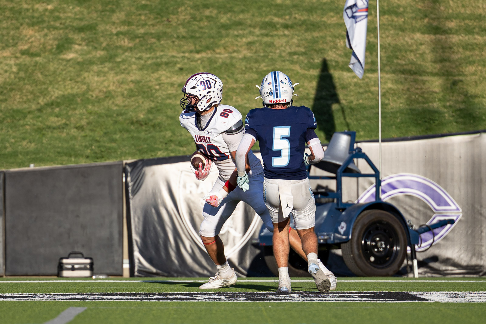 LHS-WPHS-FB-56 | Press Pass Sports Two football players contesting near the sideline: a white-uniformed receiver with the ball evades a defender in a blue jersey (#5) as they near the edge of the turf field.
