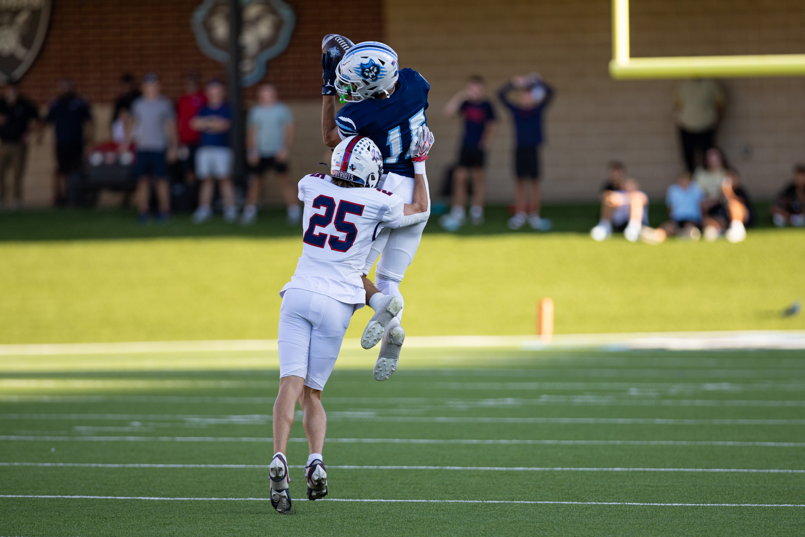 LHS-WPHS-FB-57 | Press Pass Sports Two football players leap for a catch on a grassy field, one in blue reaching for the ball while the other in white with number 25 tackles upward.