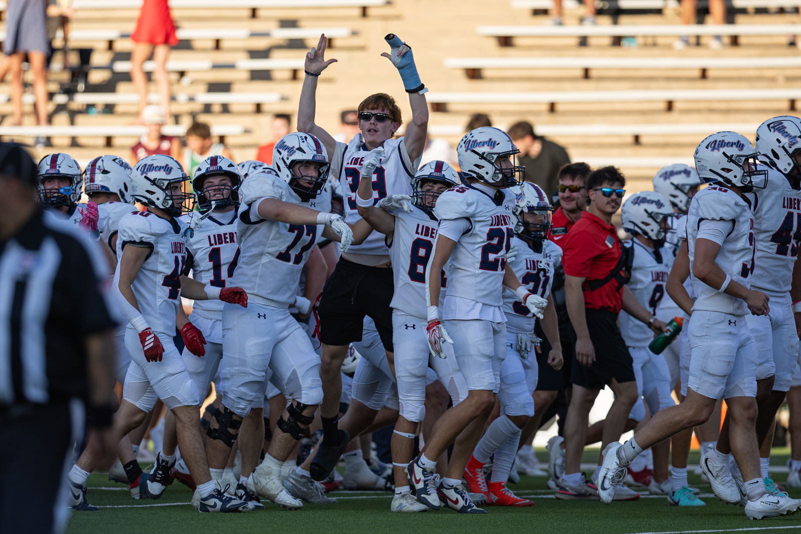 LHS-WPHS-FB-58 | Press Pass Sports Liberty High football team in white uniforms celebrates on the field, with players raising their arms amid cheers.