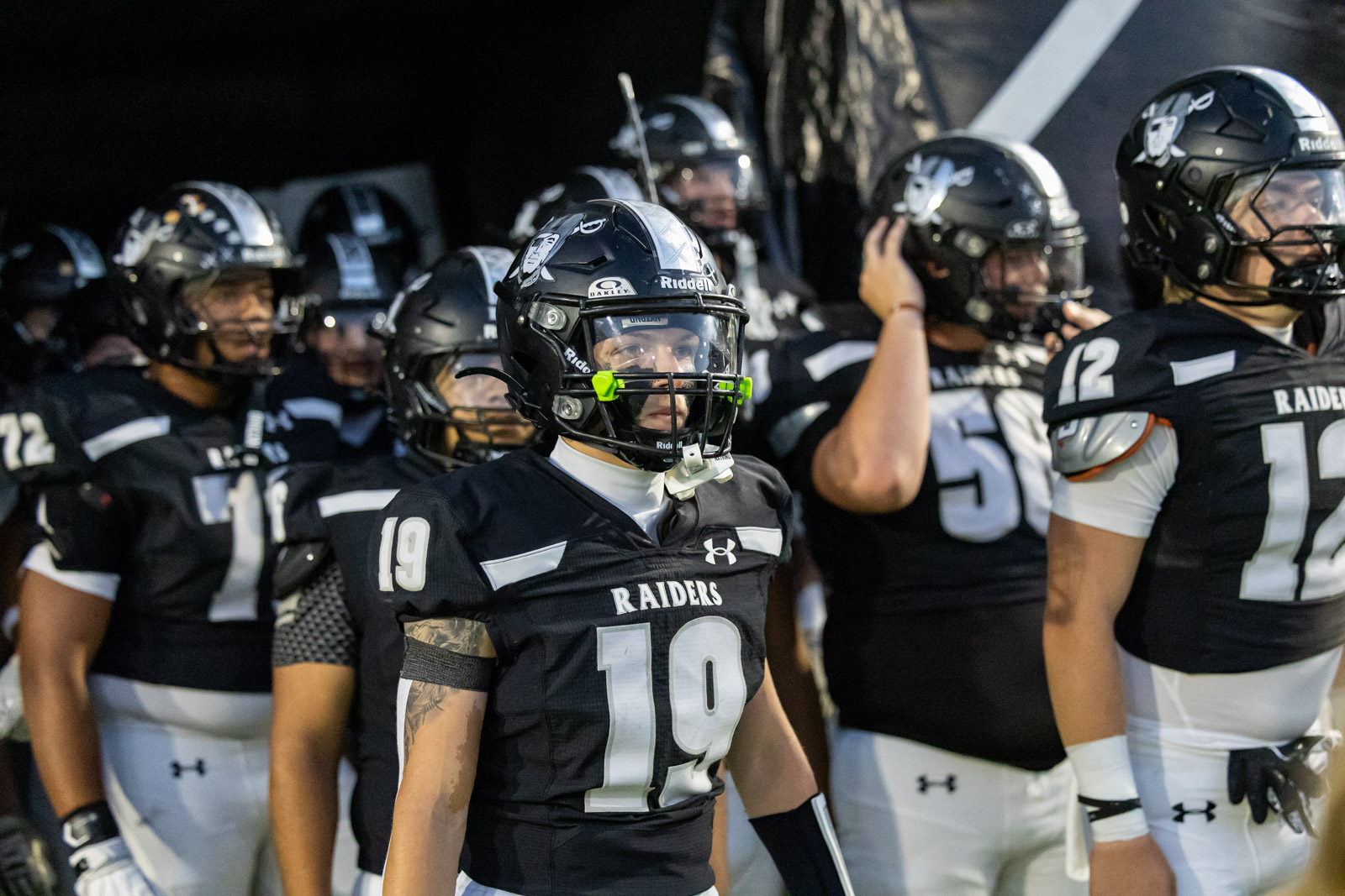 CHS-RHS-FB-2 | Press Pass Sports Football player in black Raiders uniform #19 standing in a tunnel with teammates behind him, ready for play.