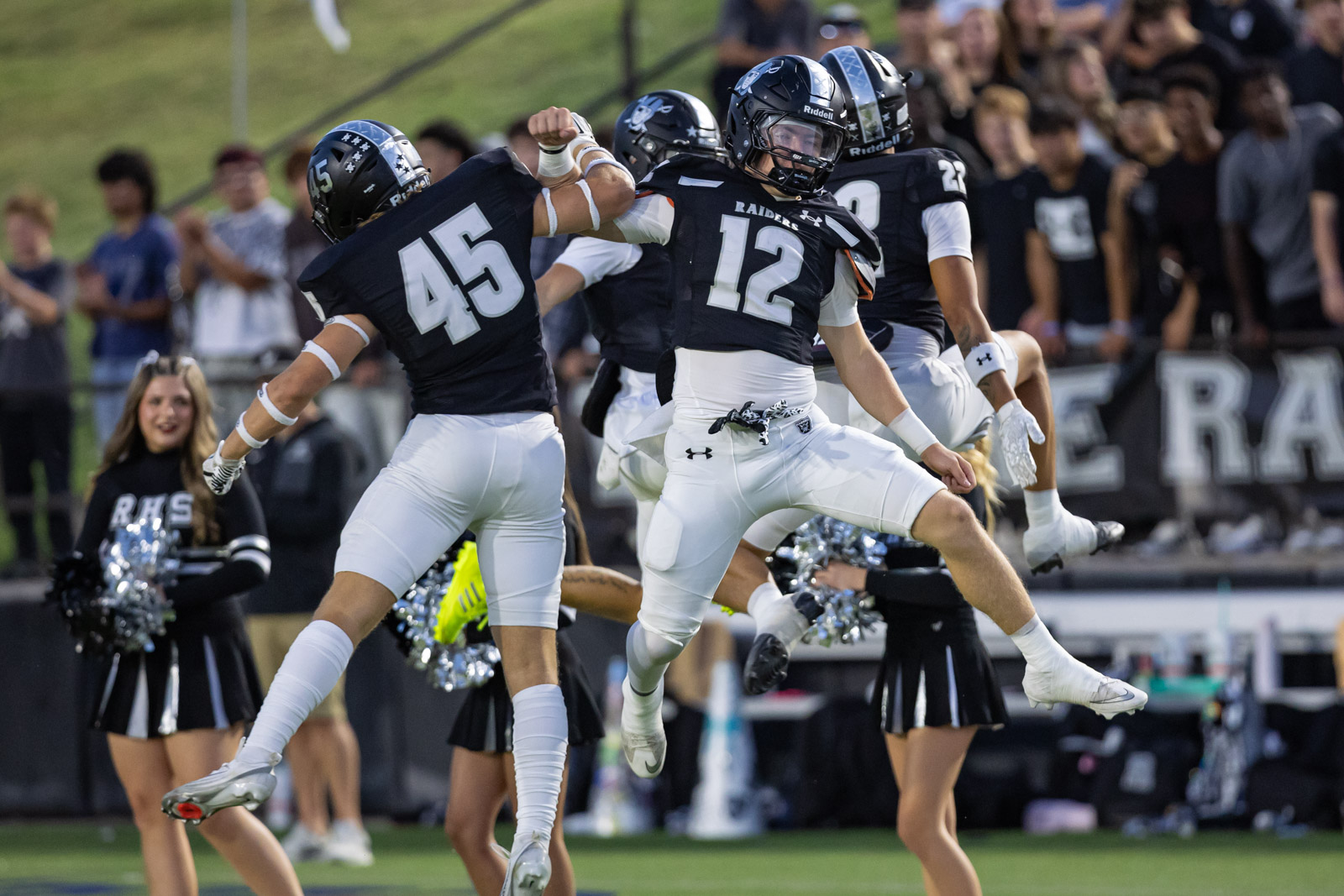 CHS-RHS-FB-7 | Press Pass Sports Football players in black uniforms jump and fist-bump in celebration on the sideline, with a cheering crowd in the stands behind them.