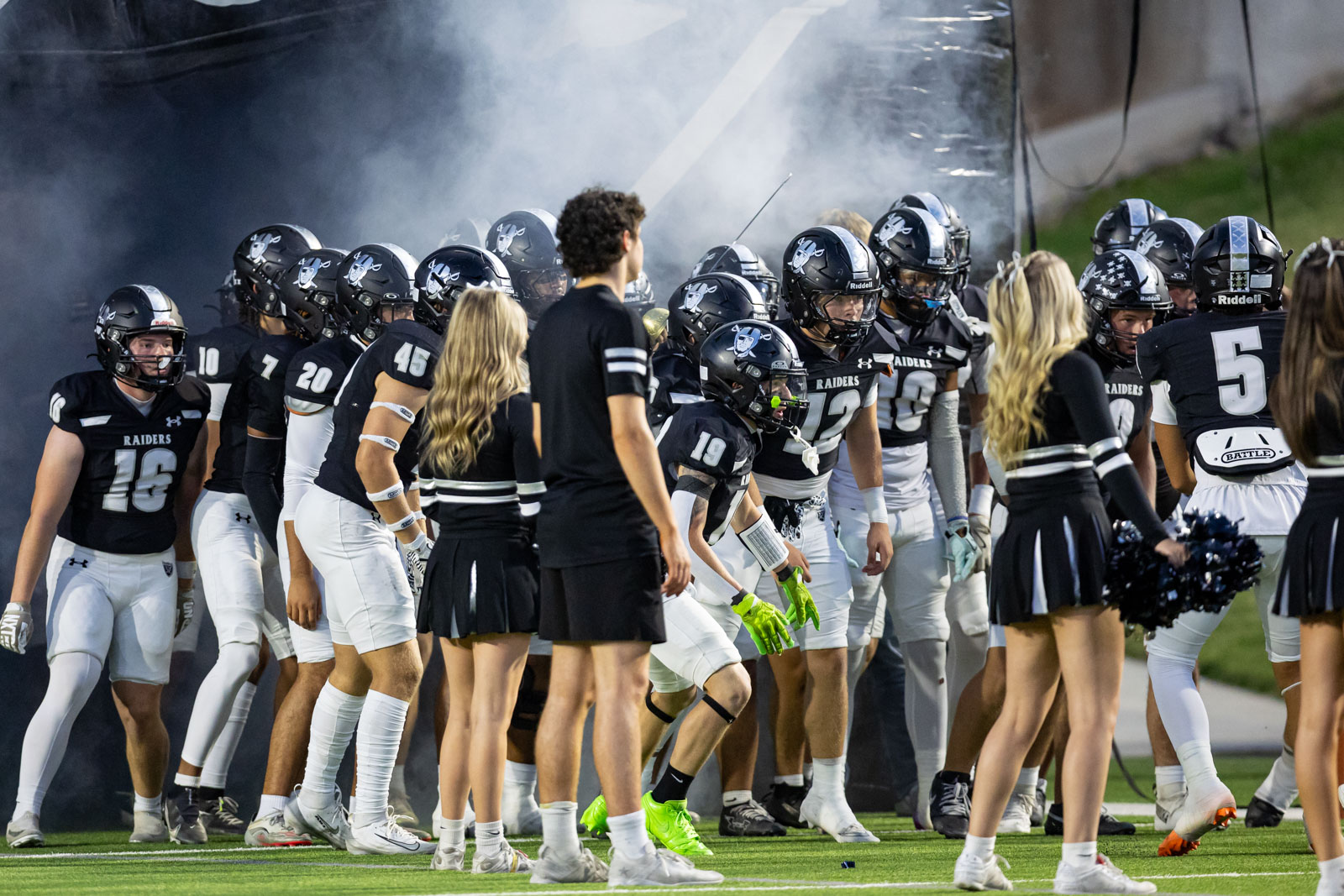 CHS-RHS-FB-10 | Press Pass Sports High school football players in black uniforms form a tunnel and huddle on the field, with cheerleaders in the foreground and smoke in the background.
