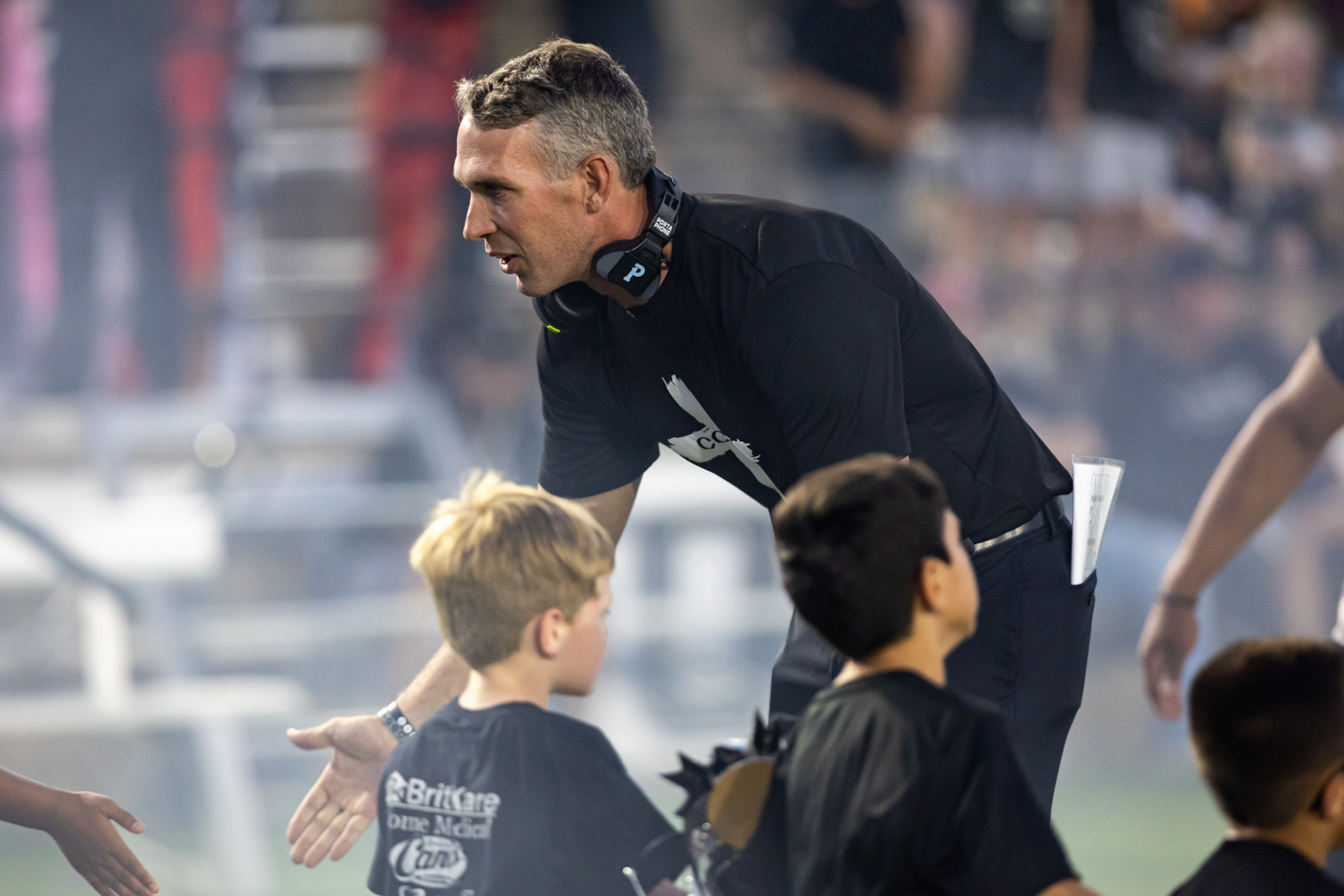 CHS-RHS-FB-11 | Press Pass Sports Coach in a black shirt bends toward young players, giving instructions during a game on a crowded field with a headset around his neck.