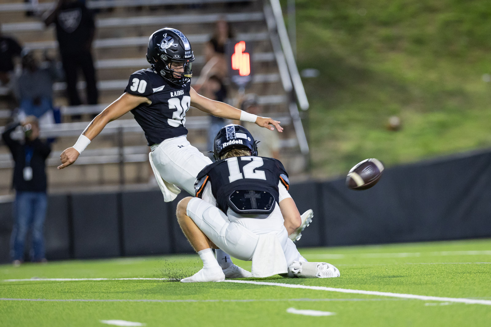 CHS-RHS-FB-13 | Press Pass Sports A football player wearing number 30 in a black Raiders uniform leaps in the air as another player on the ground reaches for the ball on a green turf field.