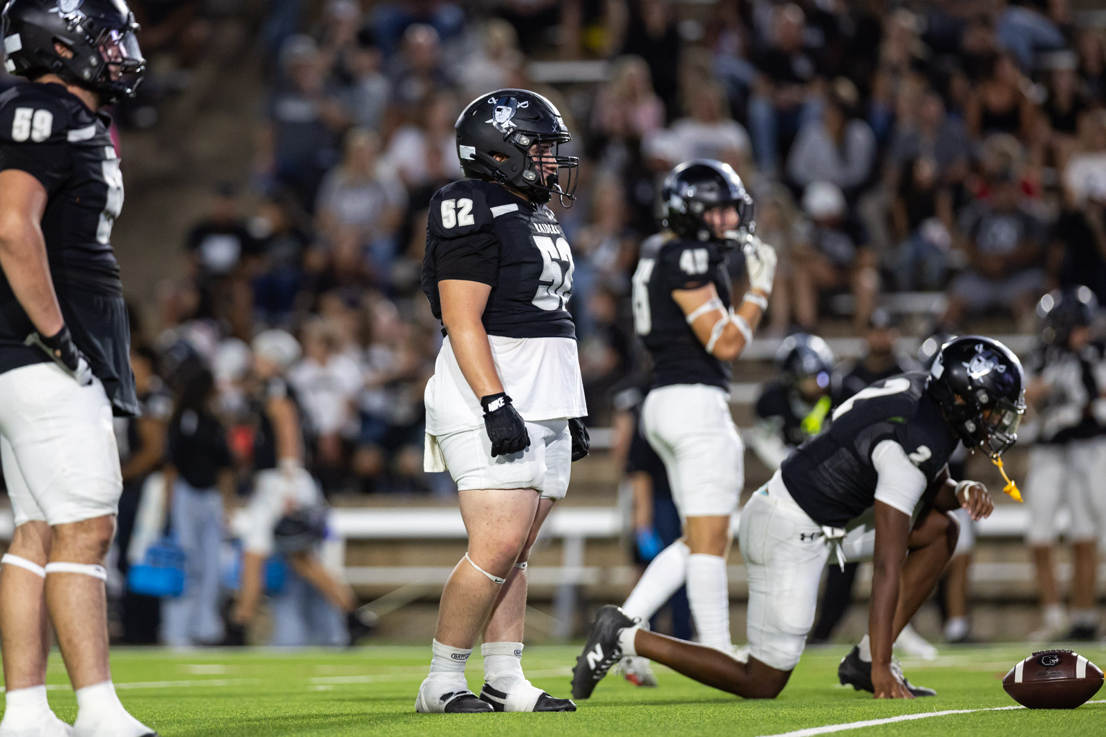 CHS-RHS-FB-14 | Press Pass Sports Football players in black jerseys on a green field during a game, central player wearing number 52 stands near the line of scrimmage as others prepare for the play.