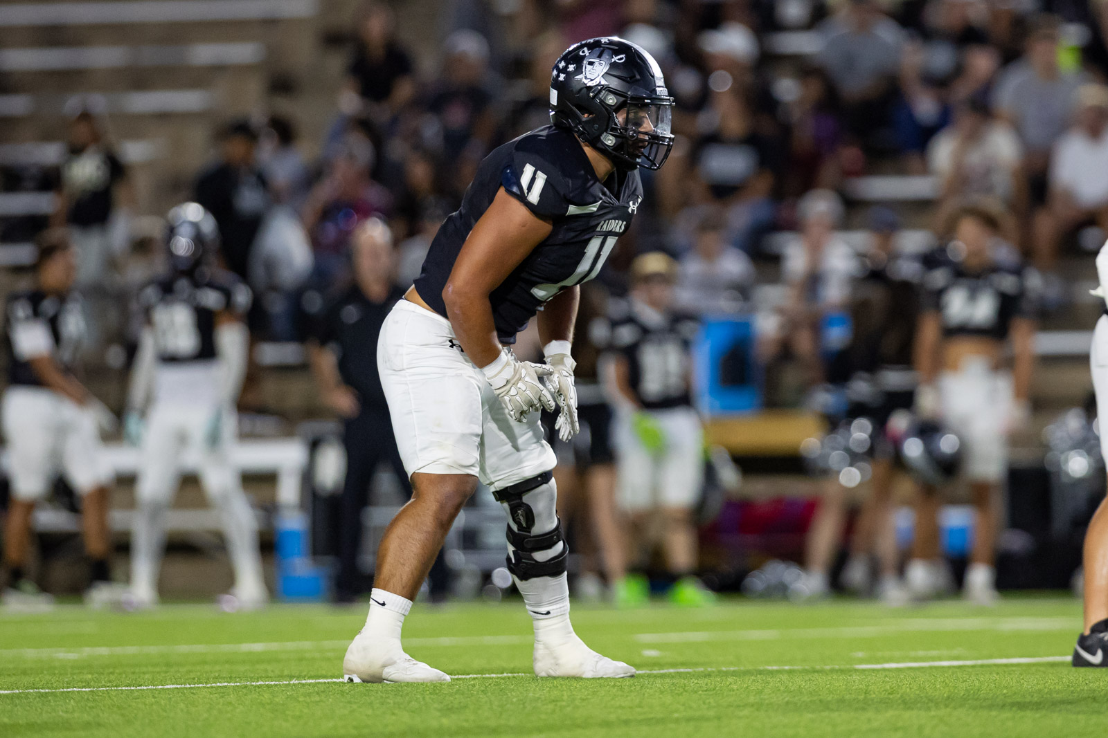 CHS-RHS-FB-15 | Press Pass Sports American football player in a black jersey with white pants crouches in a ready stance on the field, crowd blurred in the stands behind him.